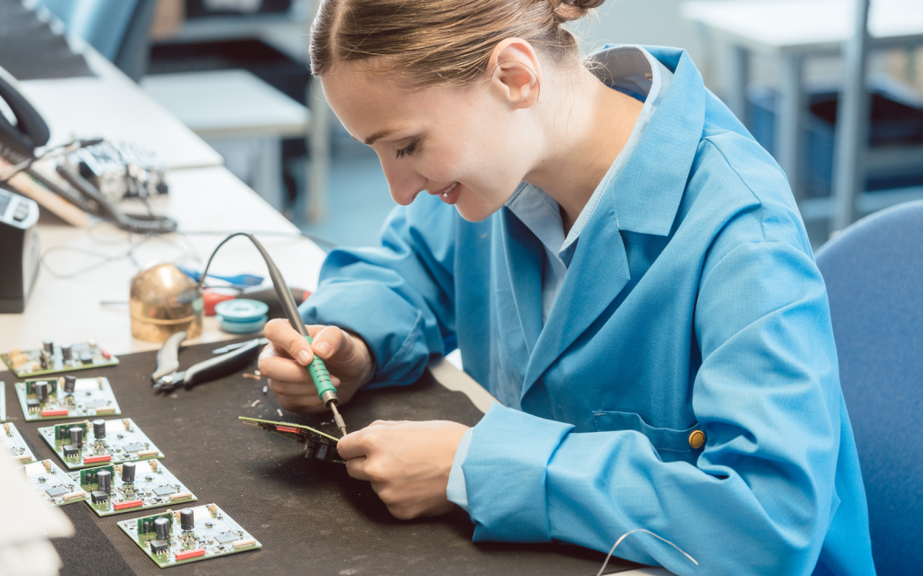 A technician in a blue lab coat carefully soldering electronic components on a circuit board at a workstation. Organized circuit boards and tools are arranged beside her, reflecting how strong inventory control systems like CyberStockroom support quality-focused production work.